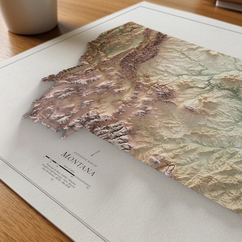 May include: A detailed, three-dimensional elevation map of Montana, featuring a textured surface with varying shades of brown, tan, and green. The map is printed on a white paper with the text "Elevation Map of Montana" and a scale indicator. The map is on a wooden surface.