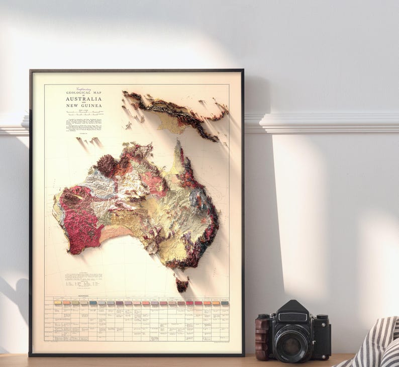 May include: A vintage geological map of Australia and New Guinea, printed in black and white with colored geological formations. The map is framed in a black frame and is hanging on a white wall.
