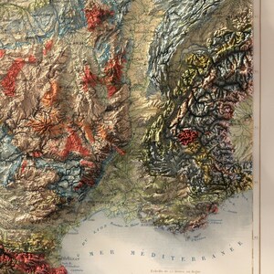 May include: A detailed, vintage relief map of a mountainous region, featuring a blend of earthy tones like beige, brown, and red. The map includes text indicating geographical features and bodies of water, with the words "MER MEDITERRANEE" visible.