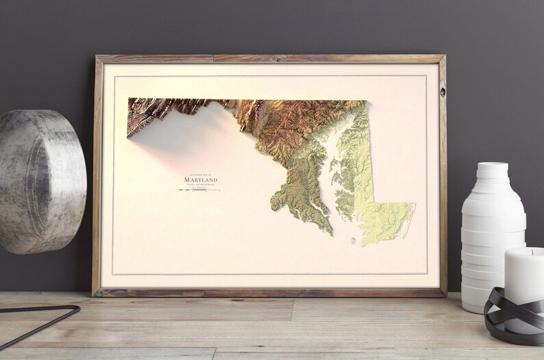 May include: A framed 3D topographic map of Maryland with a beige background. The map is in shades of brown, green, and yellow, showing the state's terrain.