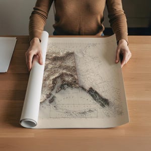 May include: A rolled-up map of Alaska is being unrolled on a wooden table. The map features a detailed relief of the Alaskan terrain in shades of brown and beige, with the Pacific Ocean and surrounding areas visible.