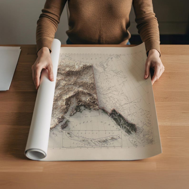 May include: A rolled-up map of Alaska is being unrolled on a wooden table. The map features a detailed relief of the Alaskan terrain in shades of brown and beige, with the Pacific Ocean and surrounding areas visible.