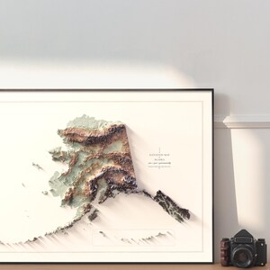 May include: Framed 3D elevation map of Alaska, featuring detailed topographical relief in shades of brown, green, and white. The map is set against a white background within a black frame. A vintage camera sits to the right of the framed map.