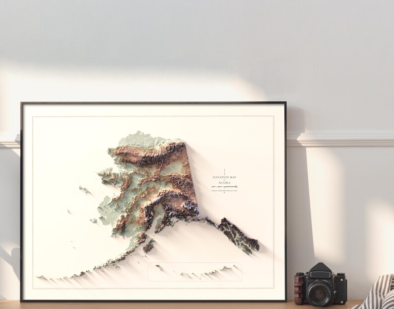 May include: Framed 3D elevation map of Alaska, featuring detailed topographical relief in shades of brown, green, and white. The map is set against a white background within a black frame. A vintage camera sits to the right of the framed map.
