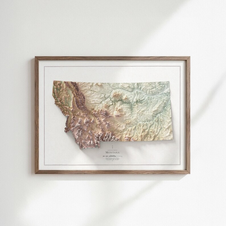 May include: A framed 3D topographical map of Montana, showcasing the state's varied terrain in shades of brown, green, and beige. The map is set against a white background and framed in a light brown wooden frame. The word "MONTANA" is printed at the bottom.