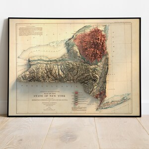 May include: Framed vintage map of New York State, featuring a 3D relief of the terrain. The map is in a black frame and has a beige background with detailed geographical features and text. The title reads "Map of the State of New York."