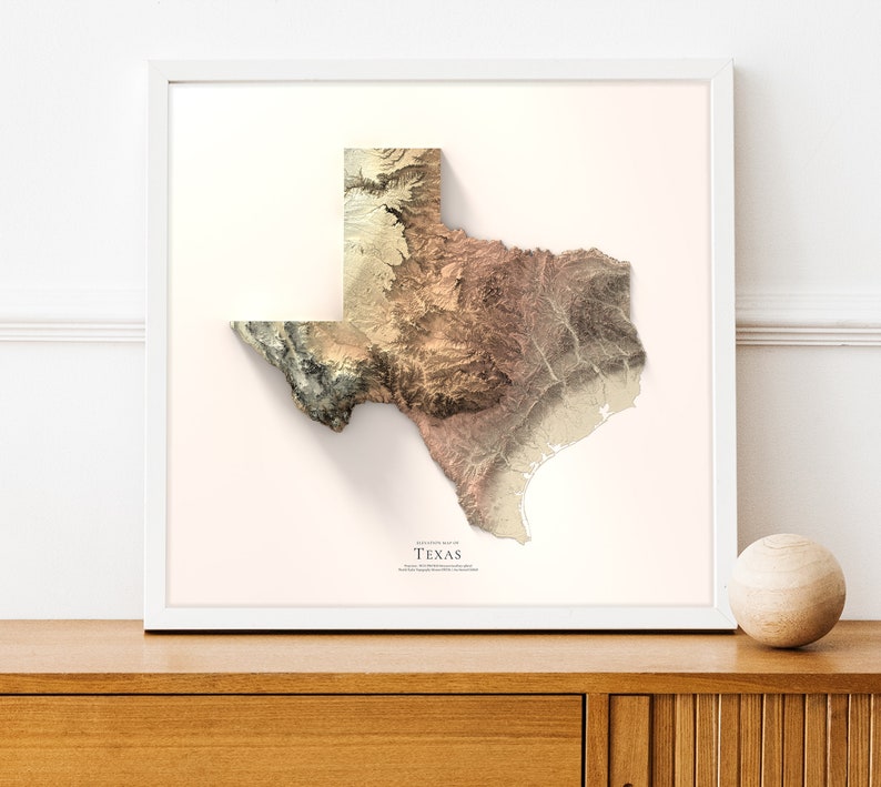 May include: A 3D topographic map of the state of Texas, printed in shades of brown, beige, and green. The map is framed in a white frame and is hanging on a white wall.