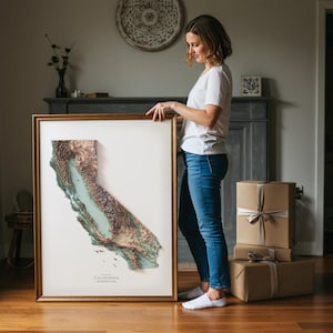 Op de afbeelding: Een ingelijste 3D-reli&euml;fkaart van Californi&euml;, met een goudkleurige lijst. De kaart toont de topografie van de staat in tinten bruin, groen en blauw. Het woord "California" staat onderaan gedrukt. Een persoon houdt de ingelijste kaart vast.