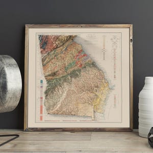 May include: A vintage map of Georgia with a detailed topographic relief showing the state's physical features. The map is printed in shades of brown, green, and blue, with a color key on the right side. The map is framed in a simple wooden frame.