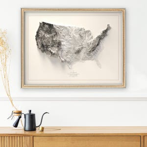 Pu&ograve; includere: Una mappa topografica in bianco e nero incorniciata degli Stati Uniti d'America. La mappa mostra l'altitudine del terreno con tonalit&agrave; pi&ugrave; scure che rappresentano altitudini pi&ugrave; elevate. La mappa &egrave; incorniciata in una cornice di legno chiaro.