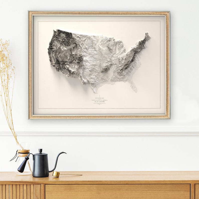 Pu&ograve; includere: Una mappa topografica in bianco e nero incorniciata degli Stati Uniti d'America. La mappa mostra l'altitudine del terreno con tonalit&agrave; pi&ugrave; scure che rappresentano altitudini pi&ugrave; elevate. La mappa &egrave; incorniciata in una cornice di legno chiaro.