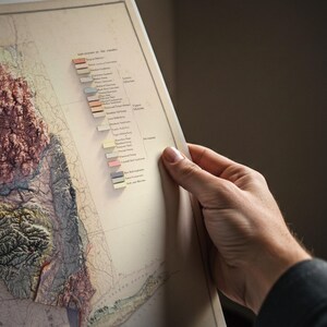 May include: A vintage map with a 3D relief of a landmass, held by a person. The map features a color-coded key indicating geological formations. The map's colors include shades of brown, green, and blue, with a detailed depiction of the terrain.