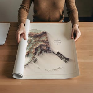 May include: A rolled-up map of Alaska, featuring a 3D elevation design in shades of brown, green, and white. The map is being unrolled on a wooden surface. The map includes the text "Elevation Map Alaska".