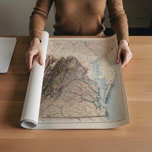 May include: A person is unrolling a vintage map on a wooden table. The map features detailed geographical information, including mountains, rivers, and coastlines. The map's color palette includes beige, brown, and blue hues, giving it a classic, aged appearance. The rolled portion of the map is white.