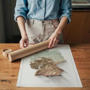 May include: A person holding a rolled-up brown paper tube next to a detailed map of Germany. The map is a 3D relief with shades of green, brown, and beige. The map is on a white background with the word "GERMANY" printed at the bottom.