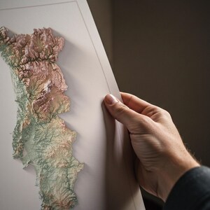 May include: A detailed, three-dimensional relief map of a landmass, showcasing varying elevations with a palette of earthy tones. The map is held by a hand, revealing its intricate topographical features and textures.