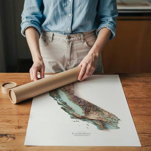 Op de afbeelding: Een persoon houdt een opgerolde bruine posterbuis vast naast een gedetailleerde topografische kaart van Californi&euml;. De kaart toont tinten groen, bruin en blauw, met het woord "CALIFORNIA" onderaan afgedrukt. De kaart ligt op een wit oppervlak.