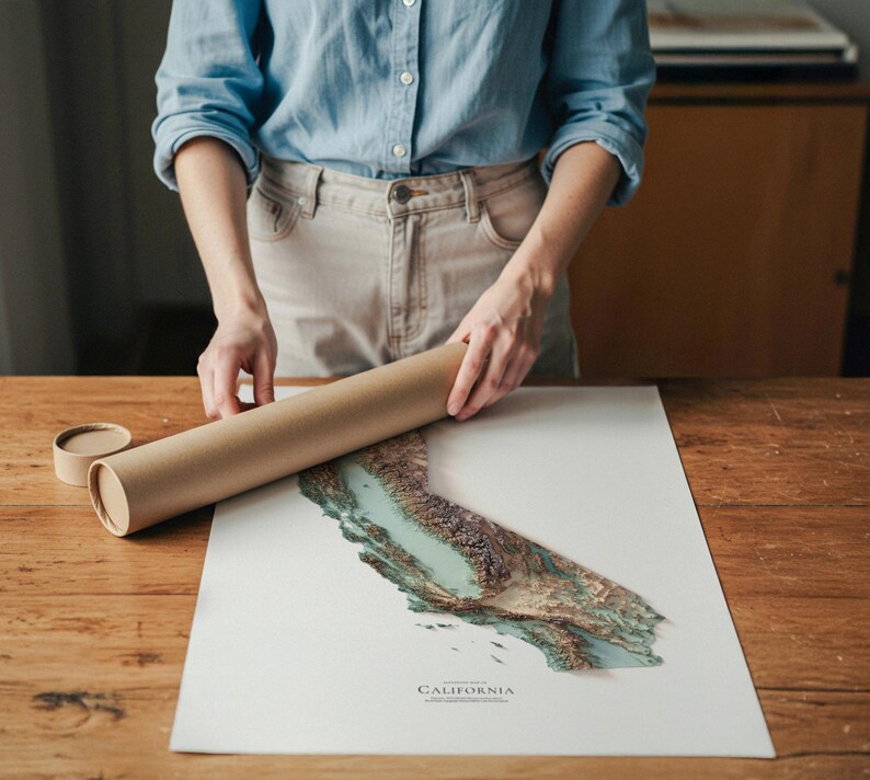 Op de afbeelding: Een persoon houdt een opgerolde bruine posterbuis vast naast een gedetailleerde topografische kaart van Californi&euml;. De kaart toont tinten groen, bruin en blauw, met het woord "CALIFORNIA" onderaan afgedrukt. De kaart ligt op een wit oppervlak.