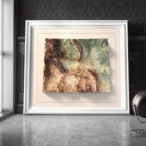 May include: A framed topographic map of Wyoming, featuring a 3D relief design with brown, green, and beige colors. The map is framed in a white frame with a cream-colored mat.
