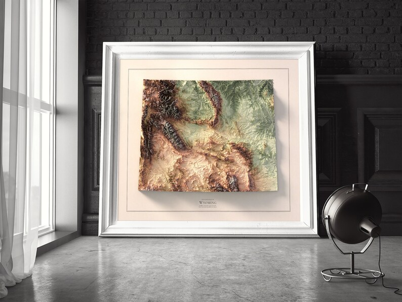 May include: A framed topographic map of Wyoming, featuring a 3D relief design with brown, green, and beige colors. The map is framed in a white frame with a cream-colored mat.
