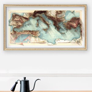 May include: A framed vintage map of the Mediterranean Sea with a 3D relief effect. The map is printed in shades of brown, blue, and green, and shows the coastline of Italy, Greece, Turkey, and other countries. The map is framed in a simple, silver frame.