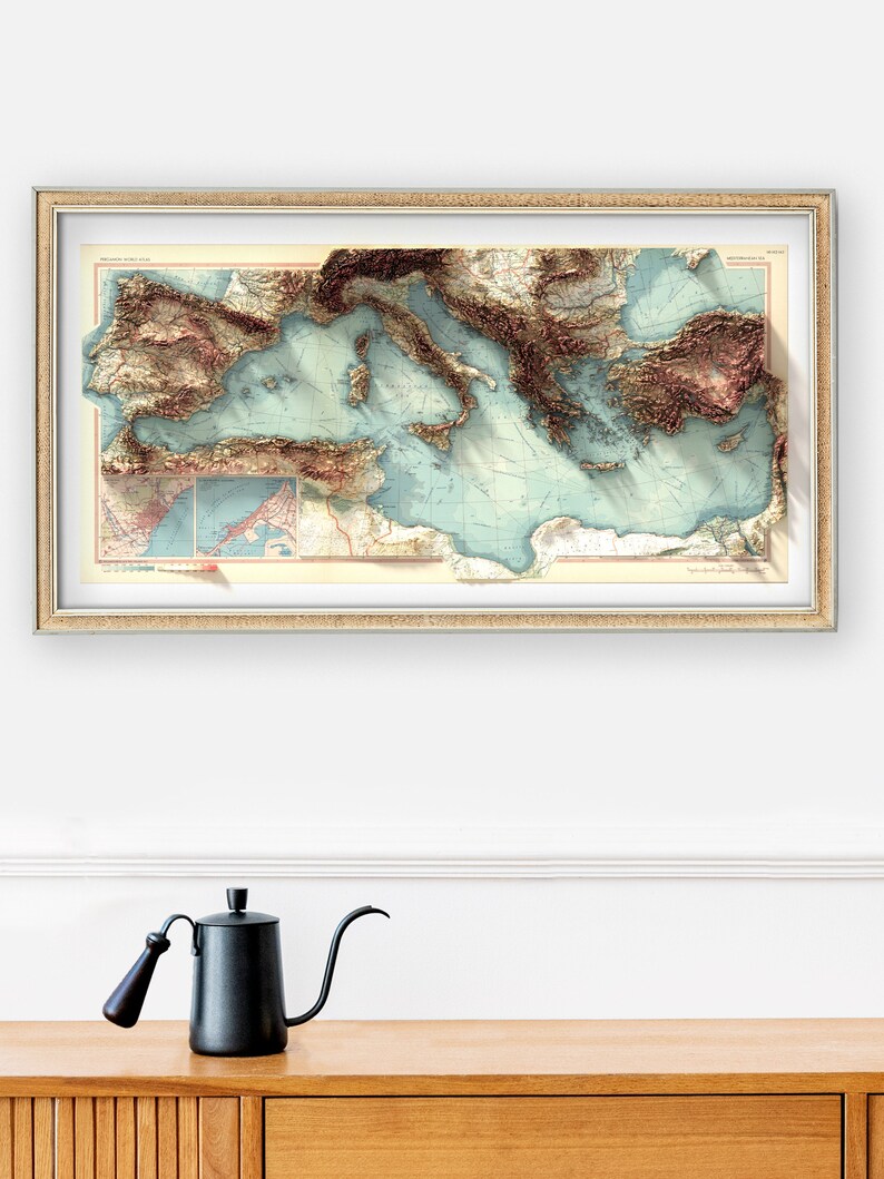 May include: A framed vintage map of the Mediterranean Sea with a 3D relief effect. The map is printed in shades of brown, blue, and green, and shows the coastline of Italy, Greece, Turkey, and other countries. The map is framed in a simple, silver frame.