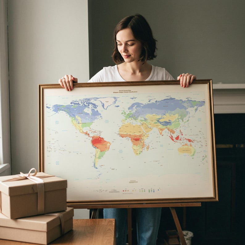 May include: A large rectangular world climate map framed in a thin gold-toned metal border. The map features a light cream background with various continents color-coded in shades of blue, green, yellow, orange, and red to represent climate zones.