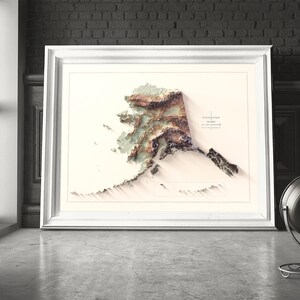May include: A framed 3D topographical map of Alaska, featuring detailed elevation contours in shades of brown, green, and white. The map is set against a neutral background within a white frame. The artwork is displayed on a concrete floor.