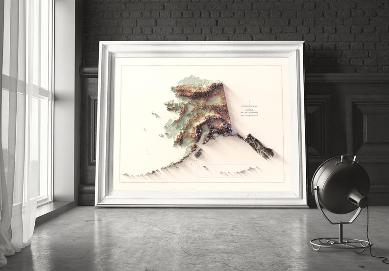 May include: A framed 3D topographical map of Alaska, featuring detailed elevation contours in shades of brown, green, and white. The map is set against a neutral background within a white frame. The artwork is displayed on a concrete floor.