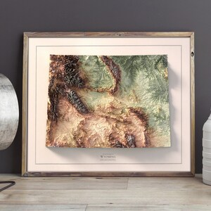 May include: A framed topographic map of Wyoming with a 3D effect. The map is printed in shades of green, brown, and beige. The frame is made of wood and has a light brown finish.