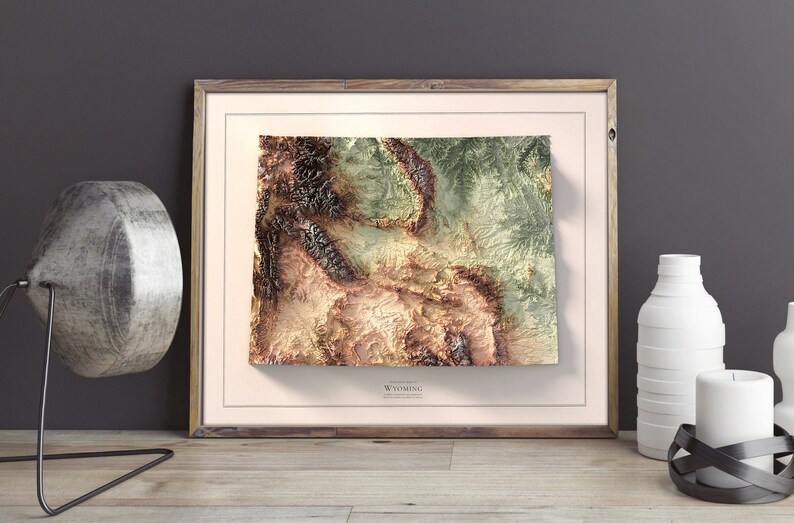 May include: A framed topographic map of Wyoming with a 3D effect. The map is printed in shades of green, brown, and beige. The frame is made of wood and has a light brown finish.