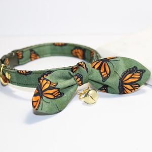 Könnte beinhalten: Ein grünes Katzenhalsband mit passender Fliege, beide mit einem Monarchfalter-Muster in Orange und Schwarz verziert. Das Halsband hat eine goldfarbene Schnalle und eine kleine Glocke. Das Set ist für Haustiere bestimmt.