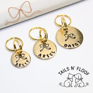May include: Three gold-colored pet ID tags with stamped names and bow designs. The tags are attached to gold key rings. The names on the tags are "Belle", "Ariel", and "Daisy". The Tails N' Floof logo is in the bottom right corner.