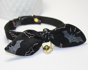 Op de afbeelding: Zwarte kattenhalsband met een roze vleermuisprint en een gouden bel. Het halsband is verstelbaar en heeft een strik.