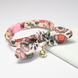 May include: A pink cat collar with a floral pattern in shades of pink, coral, and green. The collar has a bow tie and a small gold bell. The collar is designed for a cat.