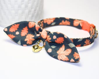 May include: A cat collar with a bow tie, featuring a dark blue fabric with orange and light green leaf patterns. The collar has a gold-colored bell and a peach-colored interior. The bow tie is in the shape of two leaves.