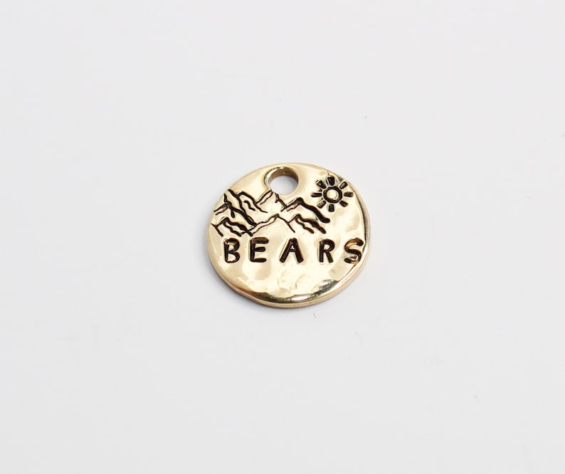 May include: Gold-colored metal charm with a mountain range design and a sun above the mountains. The charm also has the word "BEARS" engraved on it.
