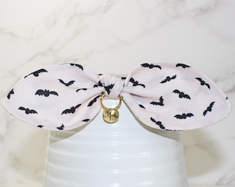 May include: A white bow tie with a black bat pattern and a gold bell. The bow tie is tied in a knot and is ready to be worn.
