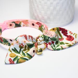 May include: A floral cat collar with a bow tie. The collar is pink and features a pattern of flowers and leaves in shades of pink, green, and red. A gold bell hangs from the bow tie.