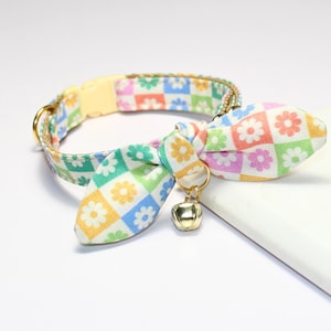 May include: A colorful cat collar with a floral bow tie. The collar features a yellow plastic buckle and a gold-colored bell. The bow tie and collar are patterned with flowers in a grid of pastel colors.