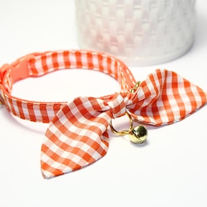 Könnte beinhalten: Ein Katzenhalsband mit rot-weißem Vichy-Muster. Das Halsband hat eine Schleife und eine kleine goldene Glocke. Es besteht aus Stoff und hat einen Kunststoffverschluss.