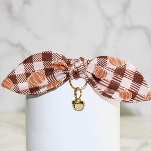 May include: A brown and white plaid dog collar with a pumpkin print and a gold bell charm.