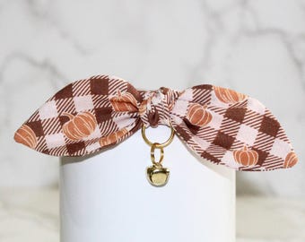 May include: A brown and white plaid dog collar with a pumpkin print and a gold bell charm.