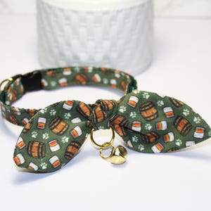 May include: A green cat collar with a bow tie featuring a pattern of orange barrels and glasses. The bow tie has a gold bell charm.