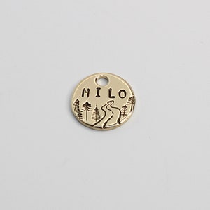 May include: Gold-colored metal dog tag with a winding path and trees design. The tag is engraved with the name "MILO".