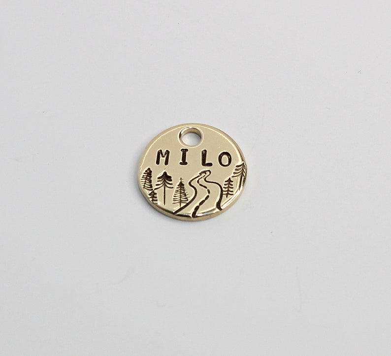 May include: Gold-colored metal dog tag with a winding path and trees design. The tag is engraved with the name "MILO".
