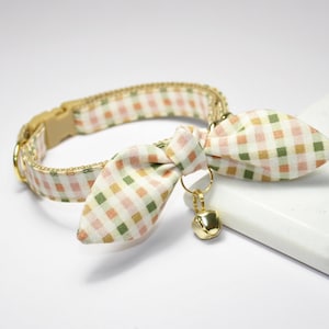 Puede incluir: Un collar para gato con pajarita, con una hebilla de plástico beige y una campana dorada. El collar y la pajarita están estampados con un diseño de cuadros pastel en tonos rosa, verde y beige.