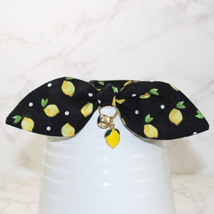 Pode incluir: Gravata borboleta preta com bolinhas brancas e estampa de limão. A gravata borboleta tem um charme de sino dourado com um pingente de limão.