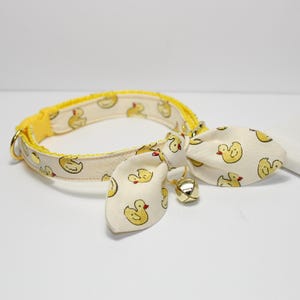 May include: A yellow and cream-colored cat collar with a duck pattern. The collar has a yellow buckle and a bow tie with a small gold bell. The duck pattern is repeated on the collar and bow tie.