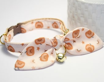 May include: A pink cat collar with a bow tie featuring a peach and white pattern of rainbows and stars. The bow tie has a gold bell attached.